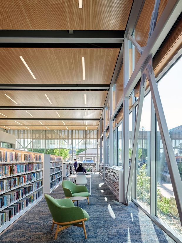 A bright contemporary library reading area with extensive floor-to-ceiling glazing, exposed structural steel bracing, timber slatted ceiling panels, linear recessed lighting, bookshelves, study tables, and green lounge chairs. The space overlooks landscaped grounds and adjacent buildings, creating an airy, daylit institutional interior.