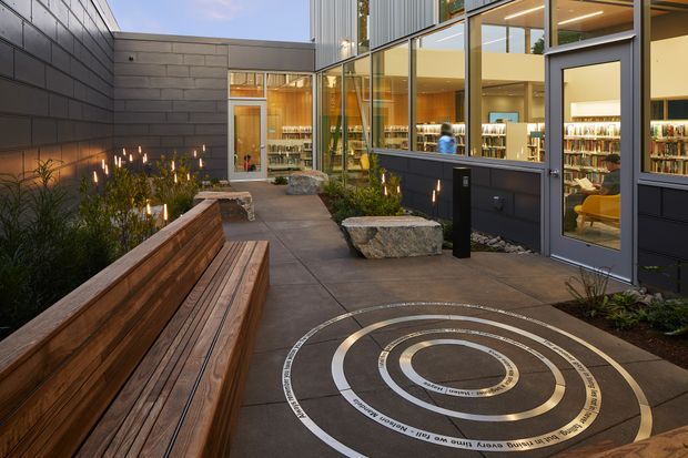 A contemporary courtyard-like outdoor forecourt beside a glazed, multi-storey building. The paved space features long built-in timber bench seating, large rough stone boulders used as seats or landscape elements, planted garden beds, a circular inlaid graphic or directional artwork in the paving, and warm architectural lighting. Full-height windows reveal a library or commercial interior with bookshelves and seating.