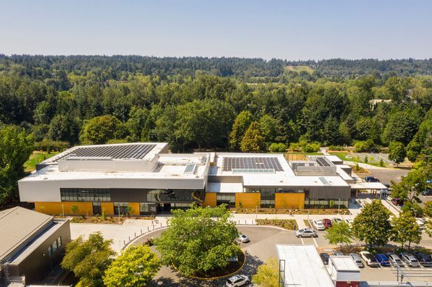 Aerial view of a large contemporary commercial building set within a leafy, wooded landscape. The low-rise structure has broad flat roof forms with extensive rooftop photovoltaic panels and skylights, a white and grey exterior, warm yellow-orange facade panels, large glazed openings, landscaped forecourt areas, and surrounding parking and access roads.