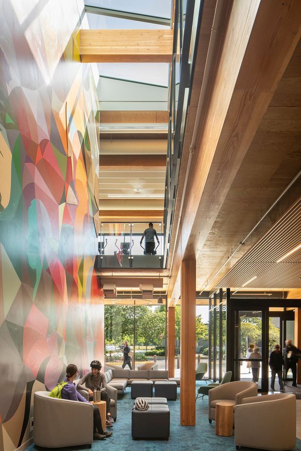 A bright, multi-level commercial interior lobby or communal lounge with exposed timber beams and columns, a glazed atrium roof, mezzanine walkway, and extensive floor-to-ceiling windows. The space includes upholstered lounge seating, small timber tables, a glass balustrade, and a large colourful geometric mural along one wall. People are using the seating and walking through the glazed entry area.