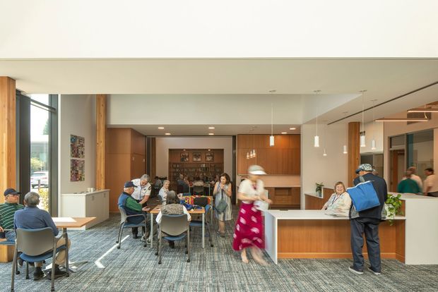 A bright contemporary communal interior, likely a reception, dining, or hospitality space, with several groups of people seated at tables and a central service or reception counter. The room features warm timber wall panelling and cabinetry, white plasterboard ceilings with recessed downlights, pendant lights, large windows, and grey patterned carpet. A glazed area and mirrored surfaces extend the sense of space, while a built-in timber counter occupies the foreground.