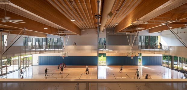 Wide interior view of a large contemporary indoor sports hall, photographed from an elevated viewing gallery. The space contains multiple basketball courts with hoops, polished timber flooring, blue padded wall panels, extensive glazing, and a striking exposed timber ceiling with linear slatted detailing and visible structural services. Bright daylight enters through the surrounding windows.
