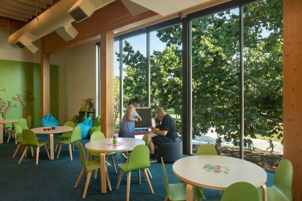 A bright, playful café or communal dining space with large floor-to-ceiling windows overlooking leafy trees. The room features round white tables, lime-green molded chairs, colorful turquoise seating, a green feature wall with mural-like graphics, dark carpet flooring, exposed warm timber ceiling panels, and visible ceiling-mounted ductwork. A few people are seated at the tables, emphasizing the space's social, family-friendly character.
