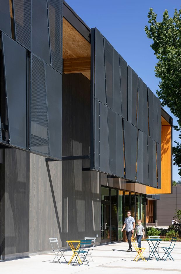 Contemporary multi-storey building exterior with a dramatic facade of dark perforated metal panels arranged in projecting and overlapping sections. Warm timber soffits and cladding contrast with the charcoal envelope, while full-height glazing opens onto a paved public terrace furnished with colourful café tables and chairs. A strong blue sky and mature tree frame the architectural composition.