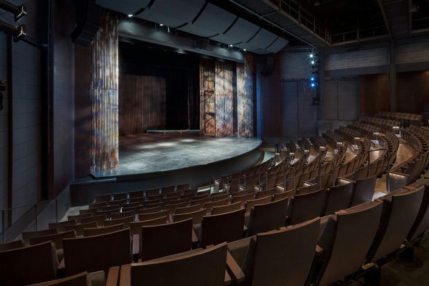 A dark, modern auditorium or theatre interior viewed from the seating area toward a broad stage. Rows of upholstered tiered seats curve around the stage, which is framed by textured vertical panels and heavy dark curtains. Stage lighting, exposed ceiling structure, and acoustic ceiling elements create a dramatic performance-venue atmosphere.