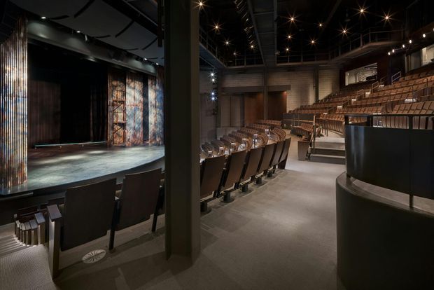 Interior view of a dark, contemporary theatre or auditorium, showing a stage at left, tiered rows of upholstered seating, a curved balcony or gallery, theatrical lighting, exposed black ceiling structure, and textured dark wall finishes.
