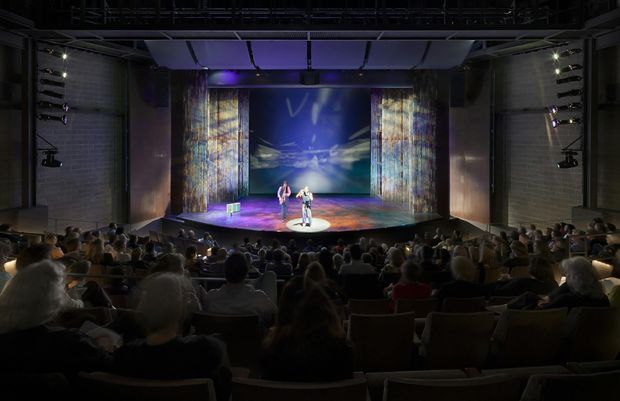 A dimly lit performance theatre viewed from the audience, with rows of seating in the foreground and a stage featuring two performers, colourful theatrical lighting, tall textured curtains or scenic panels, and a large illuminated backdrop. The venue has a contemporary black-box or adaptable theatre character with visible technical rigging and stage lighting.