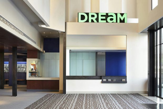 A bright, double-height commercial interior lobby or reception area with a large illuminated “DREAM” sign above a central service or reception opening. The space features polished dark timber and painted wall finishes, extensive glazing, a deep blue feature wall, a patterned carpet, and a contemporary hospitality or workplace fit-out.