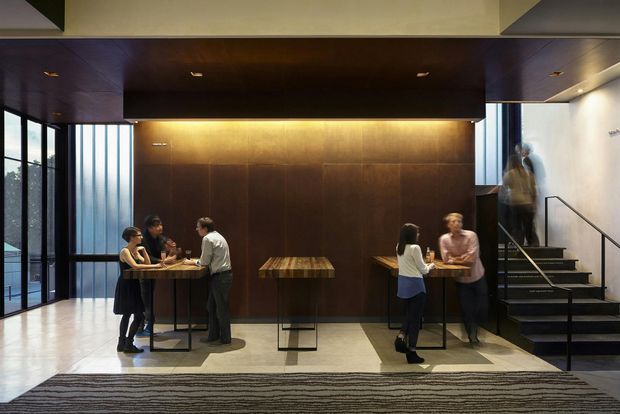 Contemporary commercial interior lobby or hospitality space with a dark timber feature wall, matching timber ceiling canopy, high bar tables, and groups of people gathered around them. Full-height glazing lines the left side, while a stairway with a metal handrail rises on the right. The space has dramatic warm lighting, polished pale flooring, and a patterned area rug.