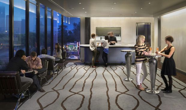 A sophisticated lounge and bar area with floor-to-ceiling glazing overlooking a city at dusk. Guests sit in lounge chairs and gather around a long stone-topped bar with high stools, while warm recessed lighting and a patterned carpet create an upscale hospitality atmosphere.