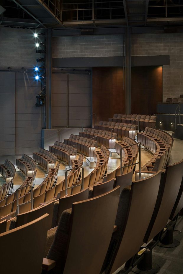 Interior view of a large auditorium or theatre with multiple curved tiers of upholstered seating, integrated aisle lighting, metal handrails, exposed structural steel, concrete block walls, and a dark industrial ceiling. Warm wood-toned seats contrast with the cool grey architectural shell.