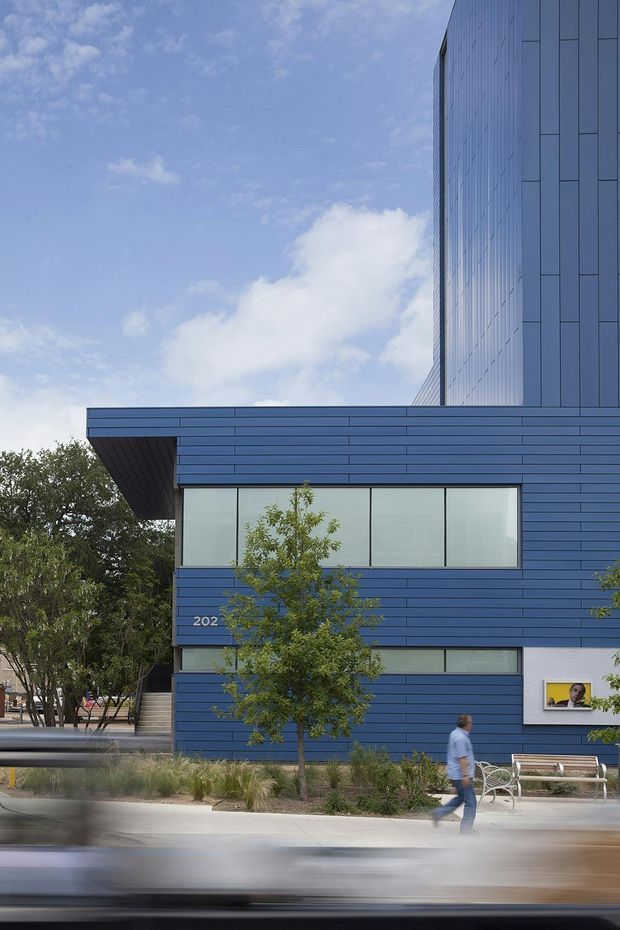 Exterior architectural photograph of a contemporary multi-storey building with a strong blue horizontal-panel facade, tall vertical upper volume, deep projecting roof edge, and large glazed windows. The building fronts a landscaped pedestrian edge with trees, grasses, benches, and a blurred passing vehicle in the foreground.