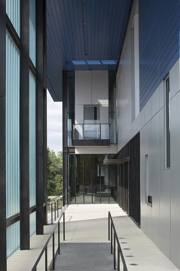 A narrow covered exterior walkway or courtyard passage between modern building volumes. The space features dark linear soffit panelling, large black-framed glazing with vertical translucent blinds on the left, pale grey metal-clad walls on the right, a concrete ramp and paving, slim black handrails, and a glass-fronted upper-level balcony or bridge. The architecture is minimalist and strongly geometric, with a skylight or roof opening bringing daylight into the passage.