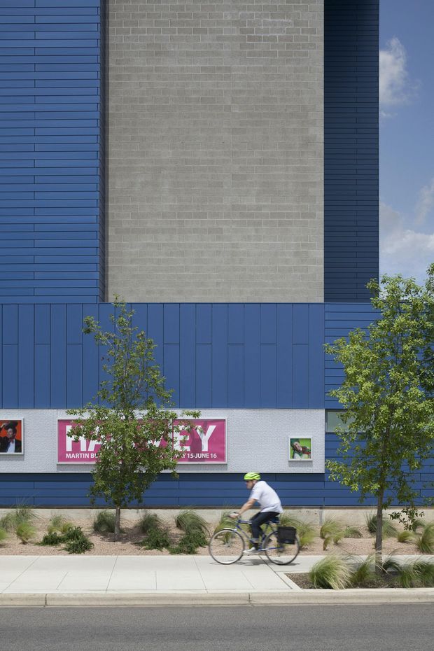 Street-level exterior view of a contemporary institutional or commercial building. The facade combines deep blue horizontal and vertical panel cladding with a tall central expanse of light grey concrete masonry blocks. Large graphic poster panels are set into the lower facade, with landscaped planting beds, a footpath, street, and cyclist in the foreground.
