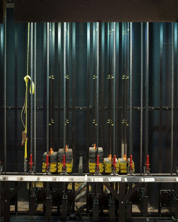 A close-up industrial interior showing a dense array of vertical ropes, cables, counterweights, pulleys, and mechanical controls mounted against a dark metal framework. The equipment appears to be stage or theatre rigging, with small labels and brightly coloured red, yellow, and orange components contrasting against black and charcoal surfaces. No conventional room furnishings are visible.