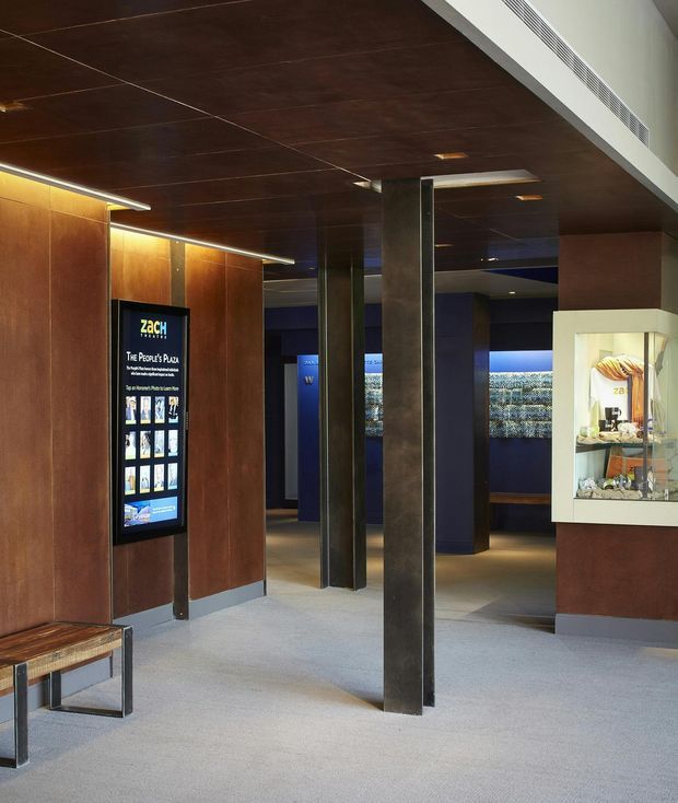 Interior view of a dark, refined commercial exhibition or visitor space with deep timber wall and ceiling panels, dark metal columns, grey carpet, recessed and linear lighting, and a glass-fronted display cabinet. A digital information screen and illuminated display wall are visible, creating a gallery or museum-like reception environment.