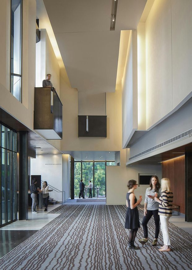 A tall, contemporary multi-level institutional or commercial interior lobby with a dramatic double-height void, mezzanine walkways, large floor-to-ceiling glazing, sculptural staircases, recessed warm lighting, dark timber wall panelling, and a patterned carpet. Several people are gathered in the spacious circulation area.