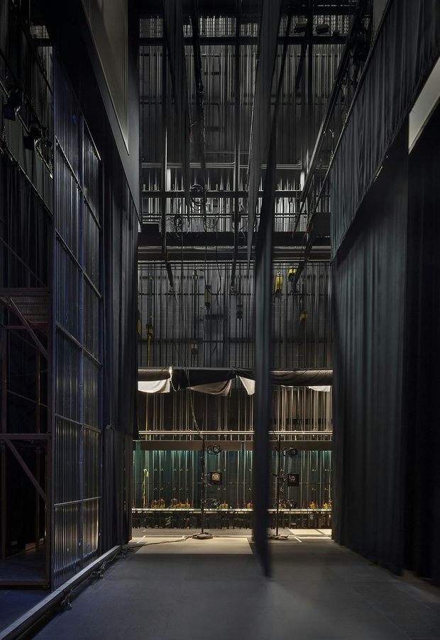 A dark, dramatic interior passage within a large industrial-style venue, looking toward a bar or service area. The space features exposed structural and mechanical services, extensive black metal framing, corrugated or ribbed metal wall surfaces, hanging fabric or mesh elements, high ceilings, and polished concrete flooring. The composition emphasizes verticality, depth, and theatrical lighting.