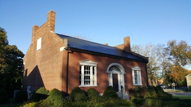 Exterior view of a small historic red-brick building with a symmetrical facade, prominent brick chimneys, white-trimmed sash windows, an arched fanlight over the central entrance, and a dark metal pitched roof. Mature landscaping and adjacent residential surroundings are visible.