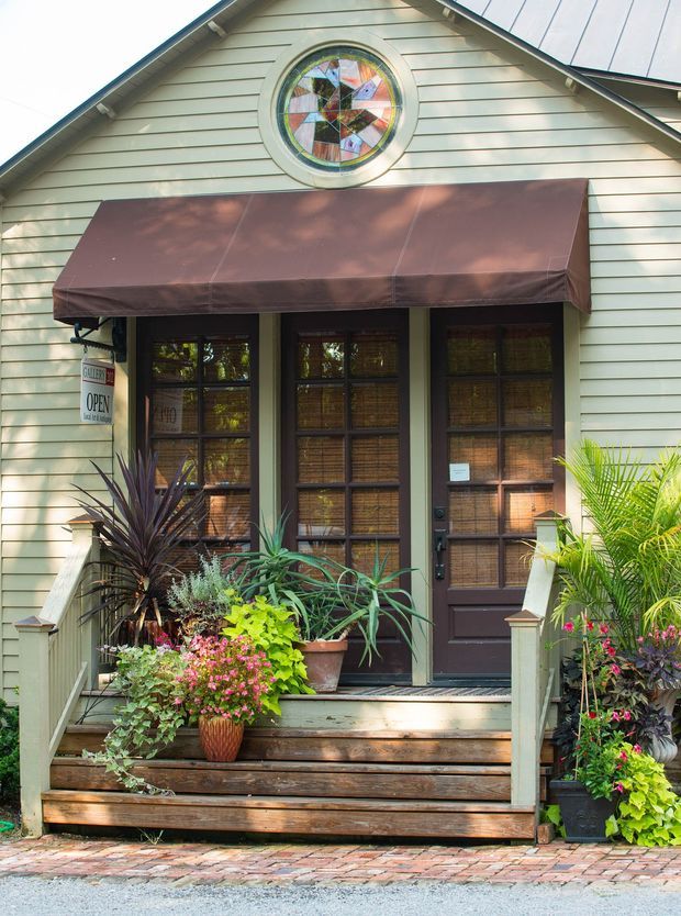 Front exterior of a small gable-roofed building with pale horizontal weatherboard siding, dark framed glazed entry doors, a brown fabric awning, decorative circular stained-glass window, timber steps and railings, and abundant potted plants flanking the entrance.