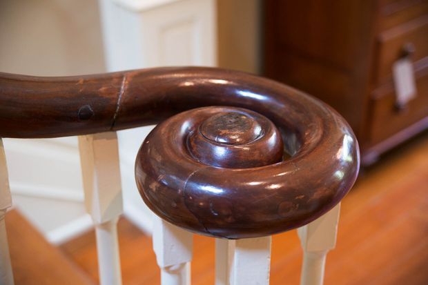 Close-up of a polished dark timber staircase handrail terminating in a large carved spiral volute. White painted balusters and traditional interior woodwork are visible behind it, with warm timber flooring and cabinetry creating a rich, heritage-style setting.