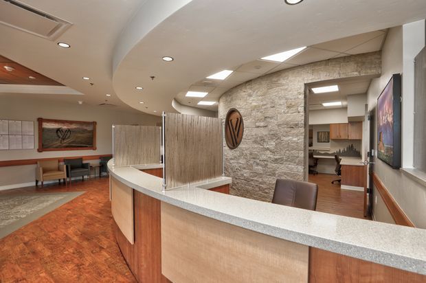 A contemporary commercial reception or waiting area viewed from behind a curved reception desk. The space features a curved light-grey benchtop, timber-look front panels, a textured stone feature wall, warm terracotta-toned flooring, wood detailing, recessed and rectangular ceiling lighting, framed artwork, seating, and a workstation visible through the opening.