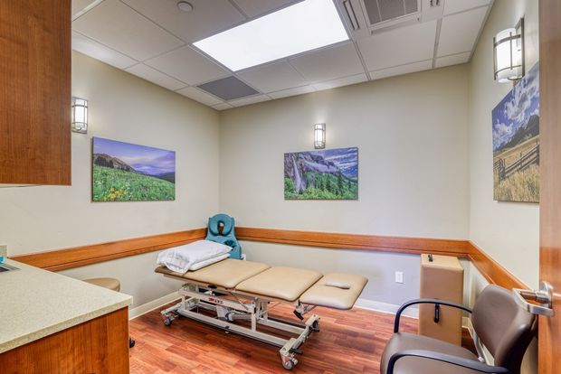 A compact clinical examination or treatment room with a padded adjustable examination table, visitor chair, built-in wood chair rail, wood-look flooring, pale walls, framed landscape artwork, and a suspended acoustic-tile ceiling with fluorescent lighting and HVAC fixtures. Fixed cabinetry and a light-coloured countertop are visible at the left.
