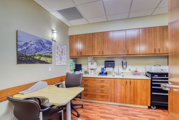 A compact, functional office or clinical workroom with extensive warm wood built-in cabinetry, a pale benchtop and sink, desk seating, printer equipment, and a small meeting table. The room has beige walls, wood-look flooring, a suspended acoustic tile ceiling, fluorescent-style wall lighting, and a mountain landscape print.