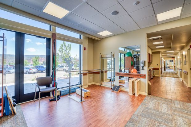 A bright institutional interior, likely a rehabilitation or wellness facility entrance and exercise area. The space has full-height glazing and a glazed entry door, wood-look flooring, carpeted circulation space, pale beige walls, a suspended acoustic tile ceiling with recessed lighting, and visible fitness or physiotherapy equipment. A long corridor extends to the right, with mirrors and handrails supporting the functional healthcare setting.