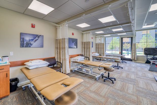 Bright clinical treatment room with multiple tan upholstered examination or therapy tables, rolling stools, privacy curtains on ceiling tracks, medical equipment, wood-toned wall trim, framed landscape artwork, and large windows. The room has a suspended acoustic tile ceiling with fluorescent panel lighting and patterned commercial carpet.