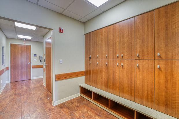 A clean institutional changing or locker room corridor with full-height warm timber locker cabinetry, integrated bench seating, wood-look flooring, pale painted walls, and a suspended acoustic tile ceiling with fluorescent lighting. A doorway and corridor extend into the background.