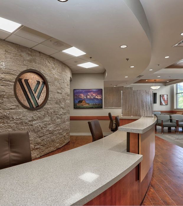 A curved reception or service counter dominates a polished commercial interior. The counter has a speckled light-grey solid-surface benchtop and dark timber-toned cabinetry, with brown leather stools and chairs. A rounded stone-clad feature wall with a large circular logo, wall-mounted television, wood-look flooring, suspended acoustic ceiling tiles, recessed lighting, and a partially screened waiting area are visible. The space has a warm, professional healthcare or office reception feel.