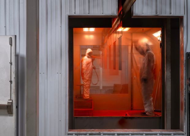 Industrial spray-painting or finishing booth viewed through a large opening, with two workers in protective coveralls inside an intensely orange-red lit enclosed workspace. The surrounding building fabric has corrugated metal wall panels, dark metal framing, and utilitarian industrial detailing.