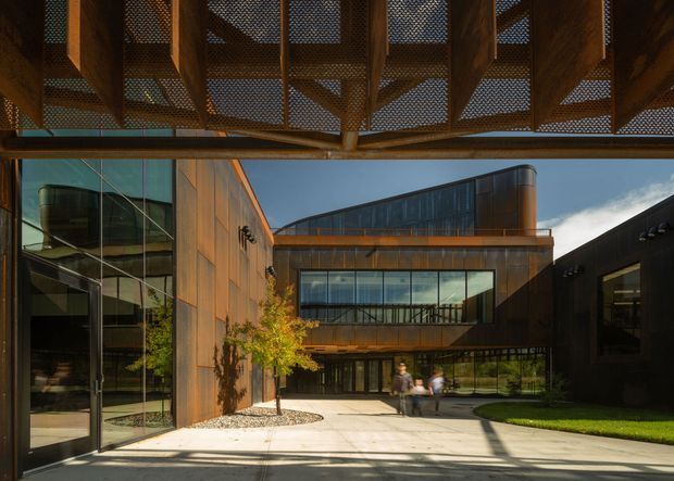 A contemporary architectural courtyard viewed from beneath a dramatic timber canopy with perforated infill panels. The courtyard is enclosed by angular buildings clad in warm weathered timber and dark metal, with extensive floor-to-ceiling glazing, a glazed bridge-like upper level, concrete paving, and restrained landscaping. The composition emphasizes strong geometric forms, exposed timber structure, and reflected light.