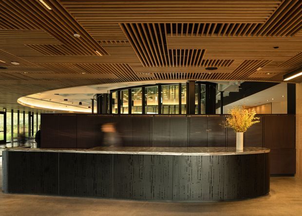 A sophisticated commercial reception area with a large curved dark perforated-metal reception desk in the foreground. Behind it is a dark timber-toned partition and glazed central room, while the ceiling features extensive geometric timber slatted panels with integrated lighting. Warm wood finishes, polished flooring, and subdued architectural lighting create a refined hospitality or corporate lobby atmosphere.