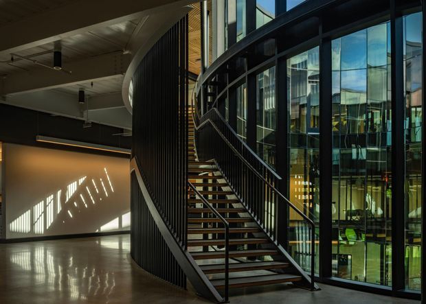 A dramatic contemporary commercial interior featuring a sculptural curved staircase with dark metal balustrades and timber stair treads. The stair rises beside a tall glazed curtain wall, revealing reflections and views into adjoining multilevel spaces. Polished concrete flooring, exposed dark ceilings, vertical screening, and strong angled sunlight create a refined, industrial atmosphere.