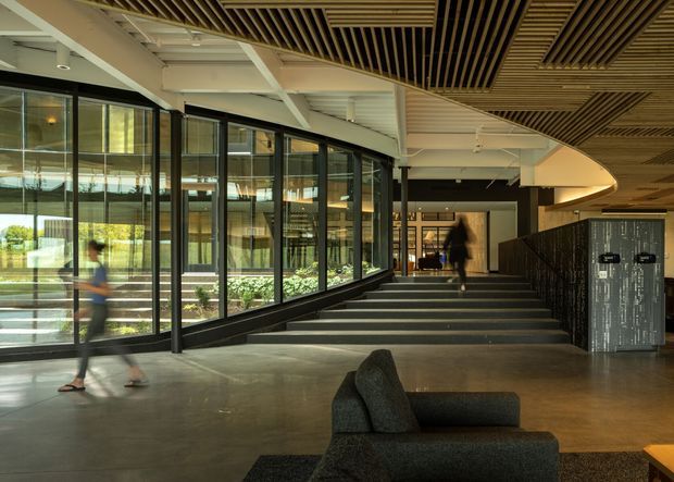 A spacious contemporary interior lobby or entry area with broad polished concrete flooring, a broad stairway, full-height black-framed glazing along one side, and a sculptural curved ceiling canopy. The ceiling combines exposed white structural beams with extensive linear timber-look acoustic slats and recessed lighting. Dark lounge seating occupies the foreground, while landscaped outdoor areas and additional building interiors are visible through the glazing.