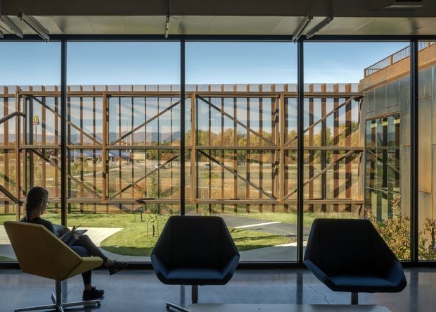A contemporary interior lounge or waiting area with large floor-to-ceiling glazed windows overlooking a landscaped outdoor space and a distinctive timber-framed structure beyond. Three modern lounge chairs sit on a smooth grey floor beneath an exposed industrial-style ceiling with visible services and linear lighting. The space is bright, open, and visually connected to the exterior.