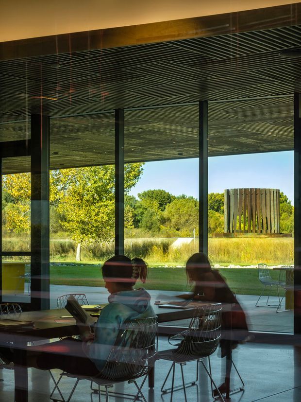 A modern glazed interior overlooking landscaped grounds, photographed through reflective floor-to-ceiling glazing. The space has a dark polished ceiling with dense linear slatted detailing, slender black structural columns, polished concrete flooring, and a meeting or dining table surrounded by wire-frame chairs. Reflections of people and furnishings appear in the glass. Outside, mature trees, lawn, and a freestanding vertical timber screen or pavilion are visible.