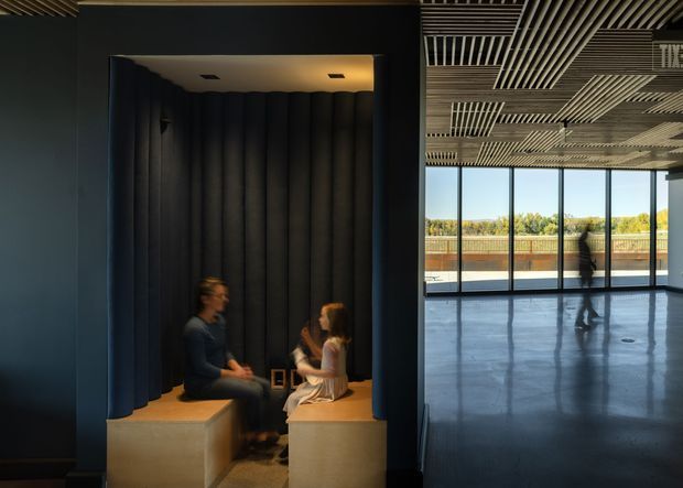 A dark, contemporary interior featuring a recessed, acoustically treated seating nook with deep vertical padded wall panels and two built-in bench seats. The nook opens onto a large, bright open-plan space with full-height glazing, a reflective polished floor, and an exposed linear slatted ceiling. Two people are seated in conversation, while an exterior terrace and vegetation are visible through the windows.