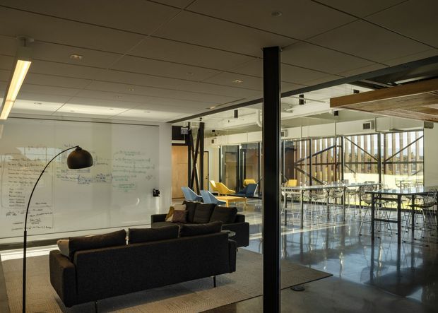 A contemporary commercial office interior with an informal lounge area in the foreground and an open dining or breakout space beyond a glass partition. The room features dark sofas, a large arched floor lamp, a wall-sized whiteboard, exposed black structural columns and framing, extensive internal glazing, polished concrete flooring, and a suspended acoustic tile ceiling. The space is bright with a functional, collaborative workplace atmosphere.