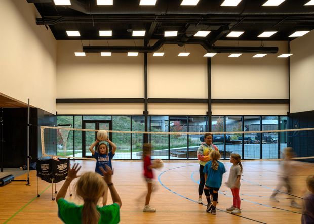 Bright indoor sports hall with a timber-finished court, basketball hoops, badminton equipment, and children playing. The space has tall pale walls, extensive floor-to-ceiling glazing, exposed black ceiling services, and evenly spaced rectangular ceiling lights.