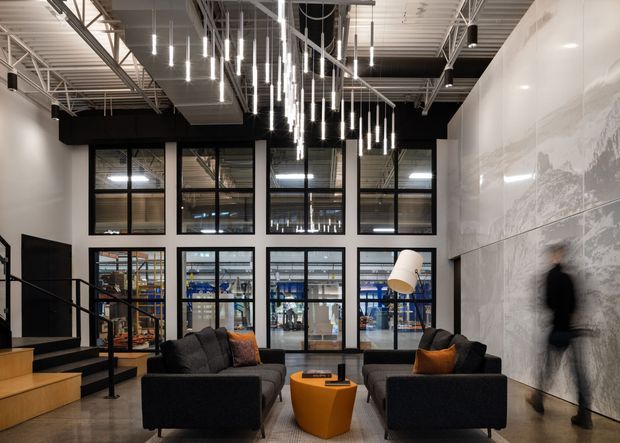 A contemporary industrial-style commercial lobby or reception area with two dark sofas arranged around a sculptural orange side table and floor lamp. The space features a high exposed services ceiling, black steel-framed glazing, polished concrete flooring, dramatic suspended linear pendant lights, timber stair elements, and a large monochrome wall mural. Warehouse or industrial activity is visible beyond the internal glazing.