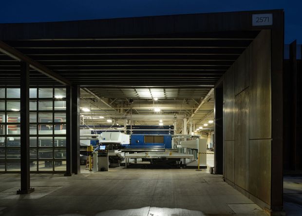 Night-time view into a large industrial workshop or manufacturing facility through a broad covered opening. The dark, rectilinear exterior portal has a deep horizontal canopy and a large metal-clad side wall, while glazed grid-framed panels on the left reveal the brightly lit interior. Industrial machinery, exposed ceiling services, and concrete paving dominate the scene.