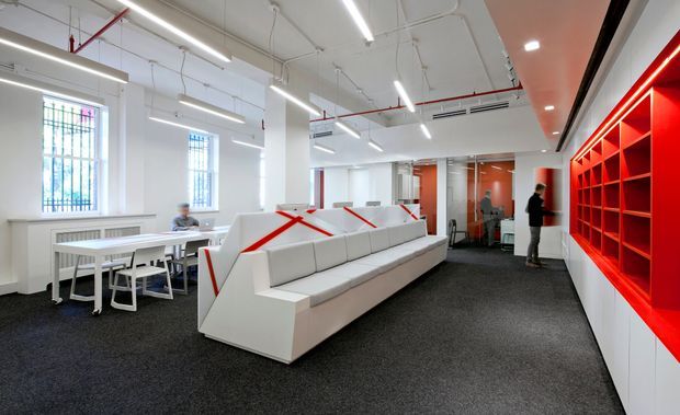 A bright contemporary commercial interior, likely an office or collaborative workspace, with exposed white services and ceiling structure, linear fluorescent lighting, tall gridded windows, white worktables, a large angular built-in white lounge bench, and a vivid red illuminated shelving/display wall. Dark carpet creates strong contrast with the predominantly white fit-out.