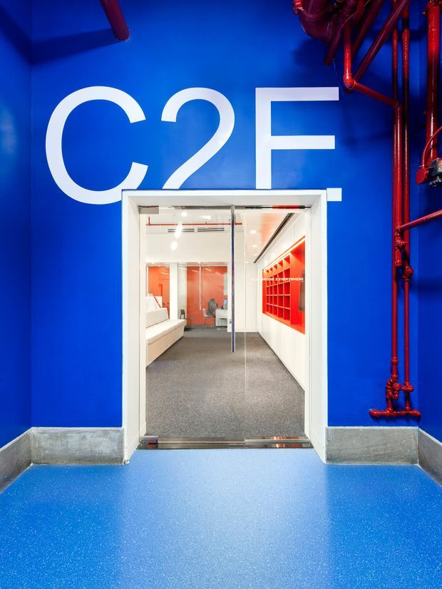 A bold commercial interior entrance with vivid blue walls and floor, large white “C2F” lettering, exposed red fire-service pipes, and a white-framed glazed doorway. Through the doorway is a bright contemporary office or commercial space with grey carpet, white seating, red feature joinery, and glass partitions.