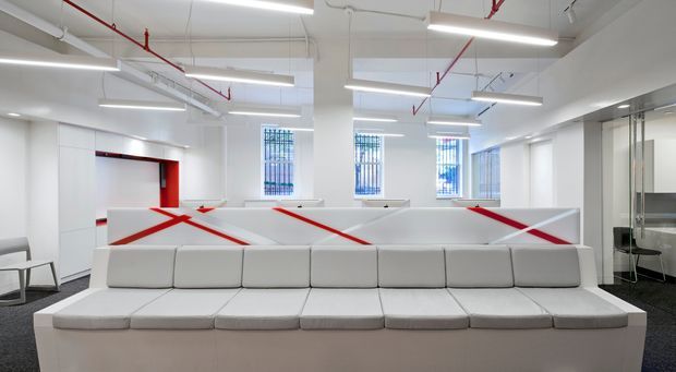 Bright open-plan commercial interior with long white upholstered modular seating in the foreground, white partitioned workstations with bold red diagonal accents, exposed ceiling services and linear fluorescent lighting. Glazed meeting or office spaces are visible to the right, with barred windows providing daylight. The setting has a clean, functional office fit-out with strong red-and-white branding.