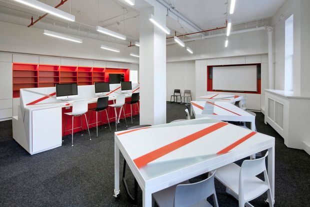 A bright, open-plan commercial workspace or studio with white walls, exposed services, fluorescent strip lighting, computer workstations, and collaborative tables. White desks and tables feature bold red-orange linear accents, complemented by red open shelving and white cabinetry. The room has a clean, functional, contemporary office fit-out.
