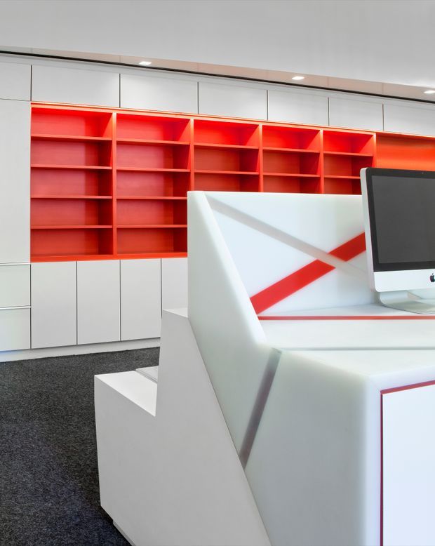 Contemporary commercial office or retail workspace featuring built-in white cabinetry with vivid red open shelving, a computer workstation, angular white desks and partitions, recessed ceiling lighting, and dark carpet. The space has a crisp geometric design with strong red-and-white contrast.