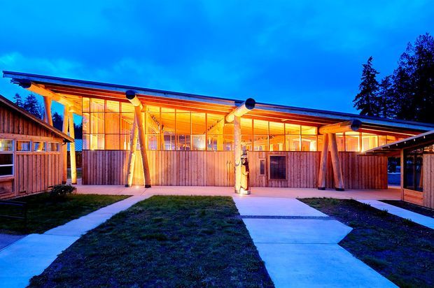 Exterior view of a low, single-storey modern building at dusk, with extensive warm-lit glazing beneath a broad shallow roof. Timber-clad privacy fences and enclosures frame concrete pedestrian paths across a landscaped courtyard, while exposed timber posts and beams support the glazed facade. A darker adjoining structure is visible at the right.